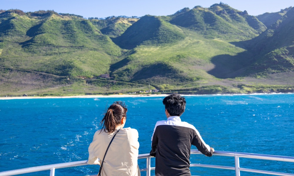 A couple standing at the bow railing, looking at the shoreline and mountains of Hawaii