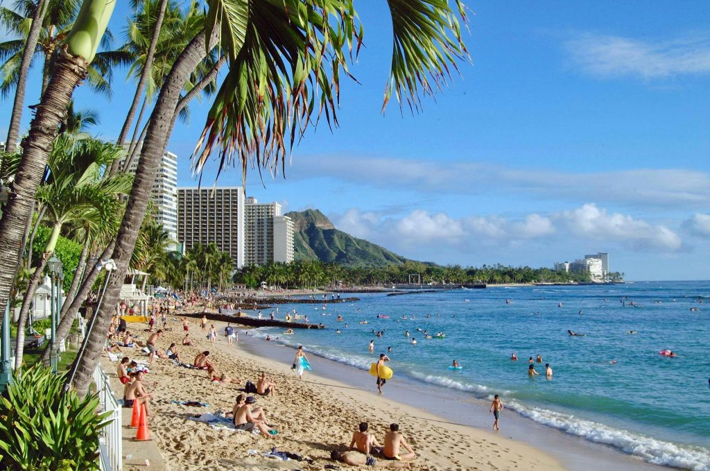 People sitting and swimming at the beach of Waikiki, along with a backdrop of mountains and tall buildings