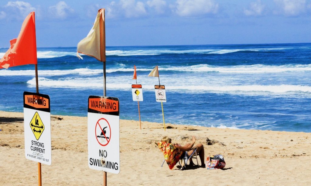 A man relaxing on a beach, near a bunch of signs, giving advisories about the conditions of the ocean